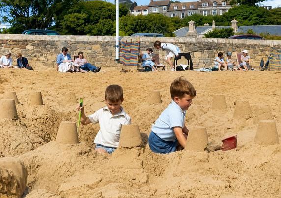 A carefree summer day finds brothers Dave and Michael Reynolds hard at work on a project of childhood importance—building sandcastles on the beach. Armed with buckets, spades, and boundless imagination, the two brothers transformed an ordinary stretch of sand into a kingdom of their own. The photograph captures the simple joys of youth, the closeness of brotherhood, and the timeless magic of childhood days spent beside the sea.